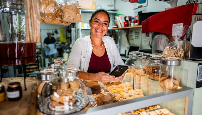 Mulher sorridente com celular atrás do balcão de uma padaria, rodeada por doces, biscoitos e be...