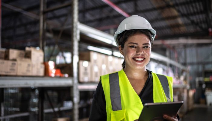 Mulher sorridente com capacete branco e colete de seguranÃ§a amarelo, segurando um tablet em um...